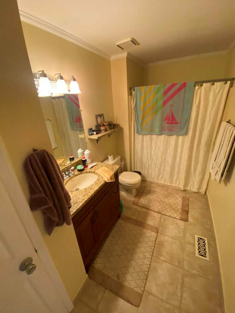 A small bathroom with a beige color theme. It features a granite countertop vanity with a sink, a mirror above, and a wall-mounted light fixture. There are two beige and brown patterned rugs on the tiled floor. The shower is in the back, with a curta