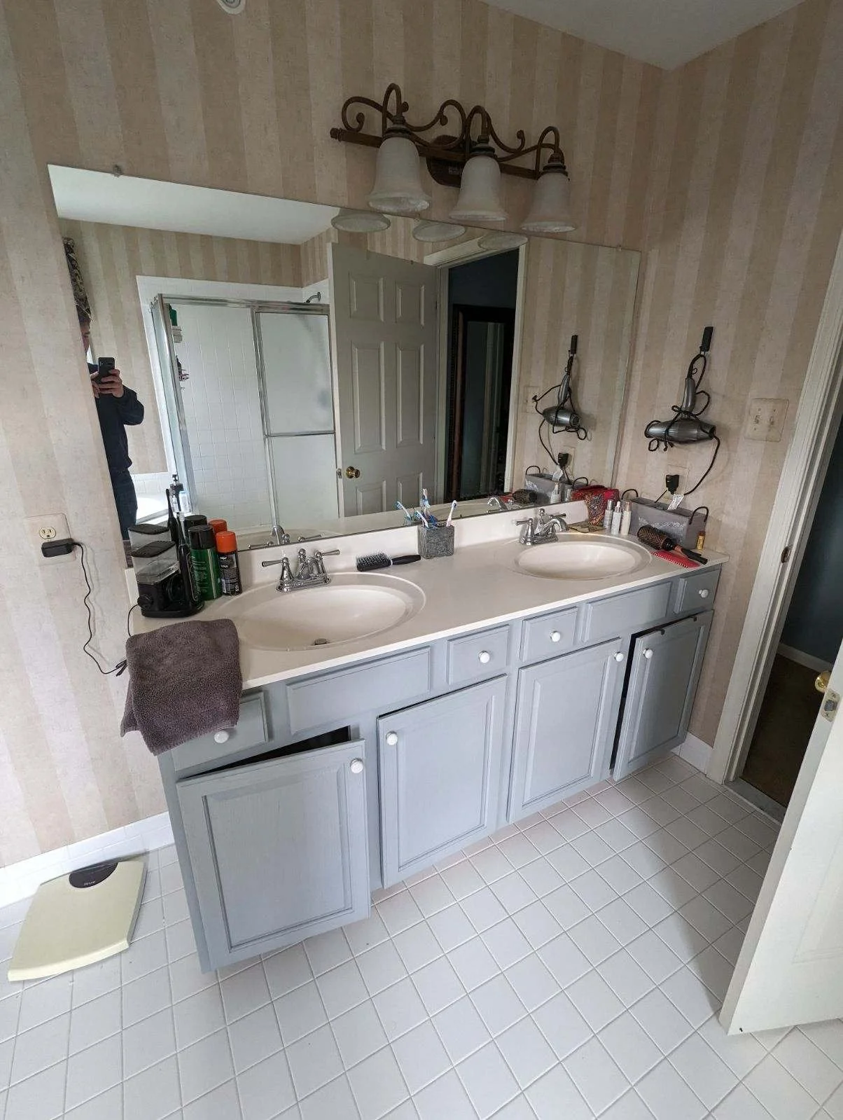 Bathroom with a large mirror, dual sinks, and light blue cabinets under a white countertop. Items such as hair dryers, toiletries, and a towel are on the counter. There is a scale on the floor and a shower with a sliding door in the background.