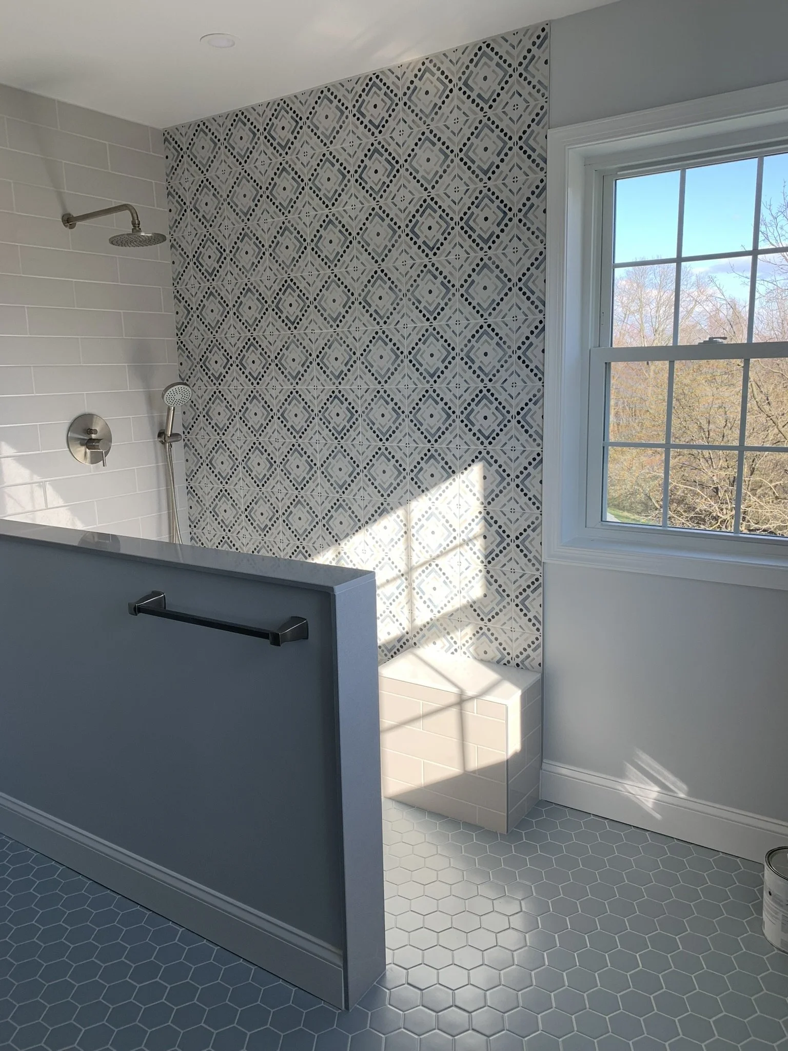 A modern walk-in shower with a patterned tile accent wall, a white subway tile wall, a large window with a view of trees, and a gray privacy wall with a black towel bar.