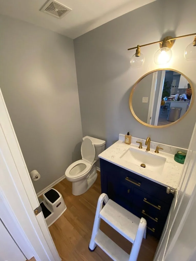 Small bathroom with light gray walls, a white toilet, a navy blue vanity with gold hardware, a white marble countertop, a round mirror with a gold frame, and three globe light fixtures above the mirror.