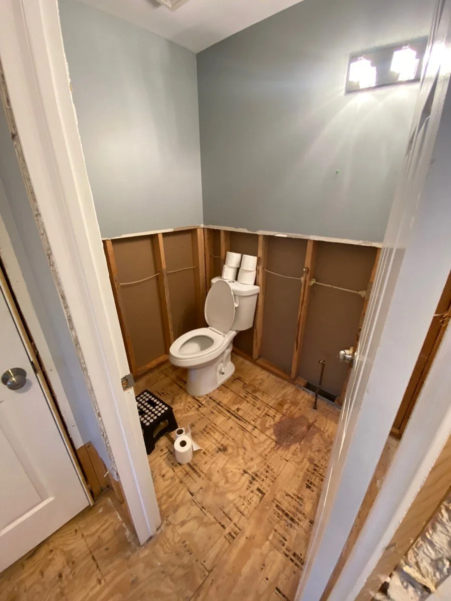 A small bathroom with a toilet, two rolls of toilet paper on top of the toilet tank, a small step stool, and a roll of toilet paper on the floor. The walls are partially unfinished, revealing wooden framing, and the floor is plywood. There is a gray 