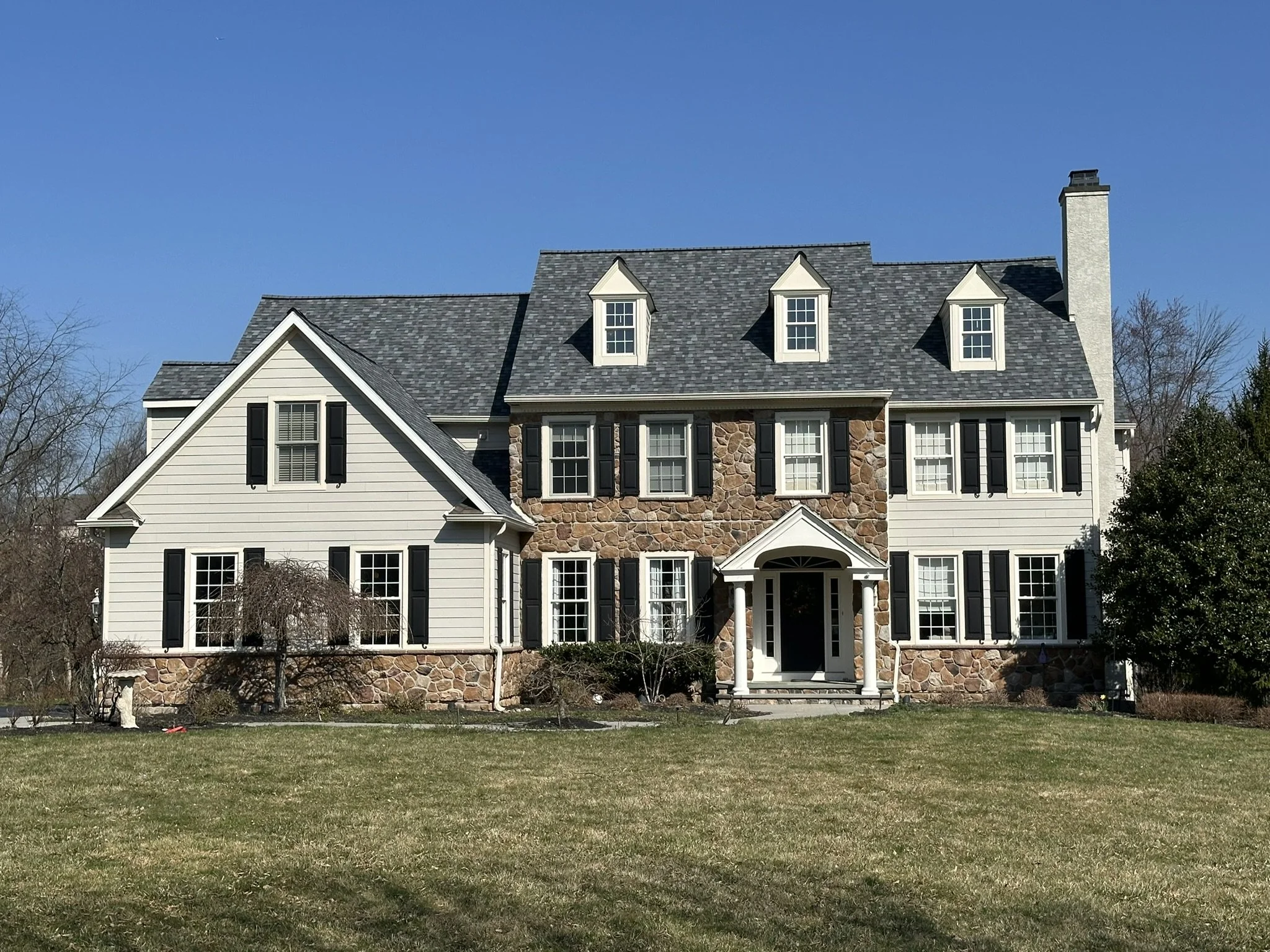 Large two-story house with white siding, black shutters, and a stone facade, under a clear blue sky.