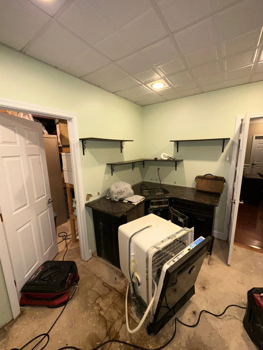 A small, cluttered kitchen space with a countertop, black cabinets, and two wall shelves. There is an open door leading to what appears to be a laundry or storage room. Various tools and appliances, including a portable air conditioner or dehumidifie