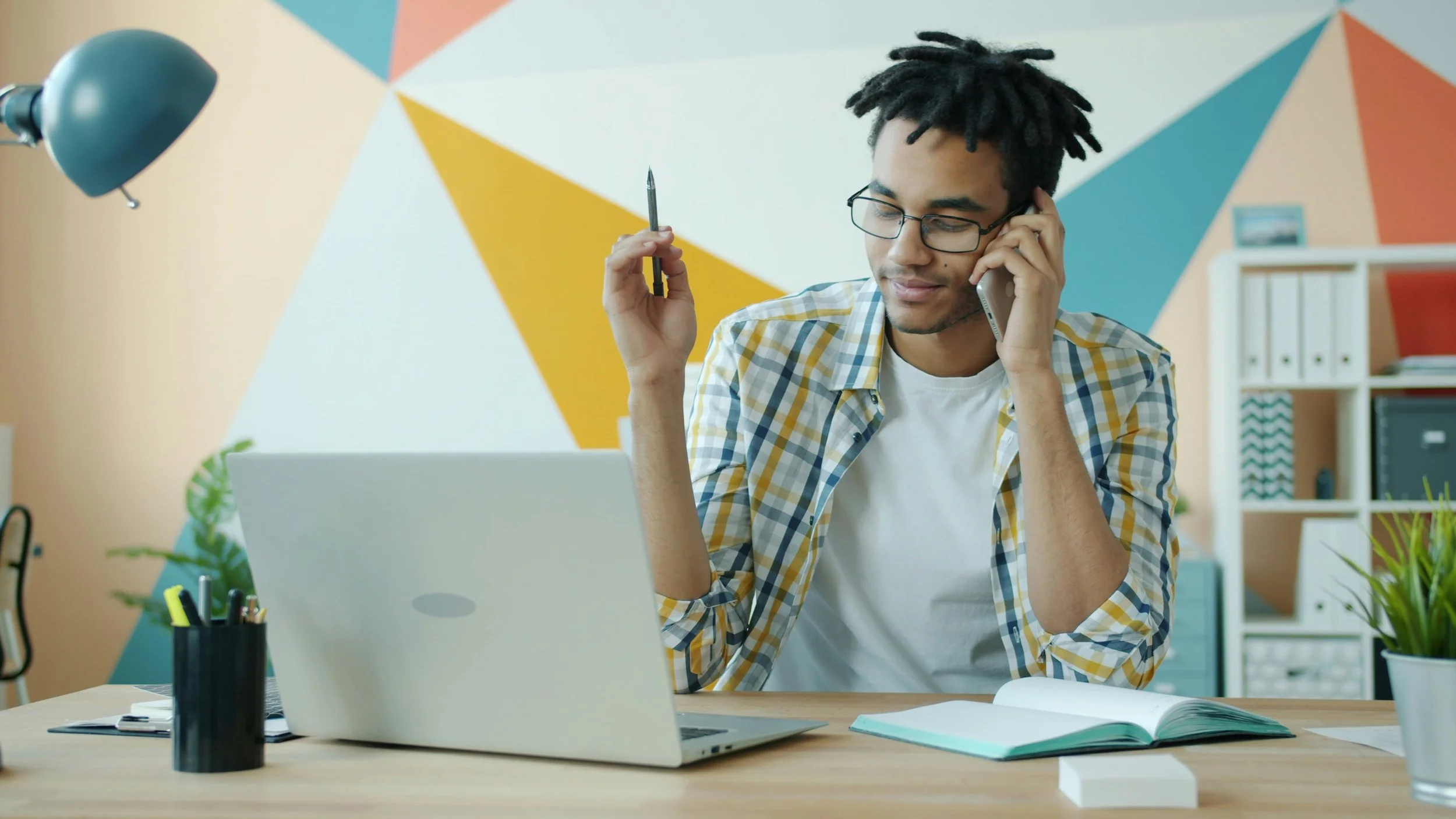 A man takes notes during a phone call at his desk, laptop and notebook open in front of him.