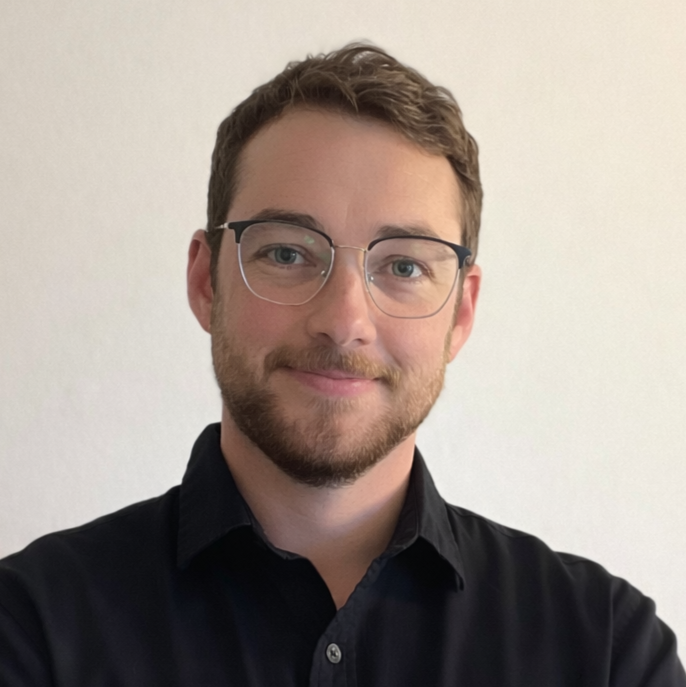 A young man with glasses and a beard, wearing a black shirt, smiling and crossing his arms against a plain white wall.