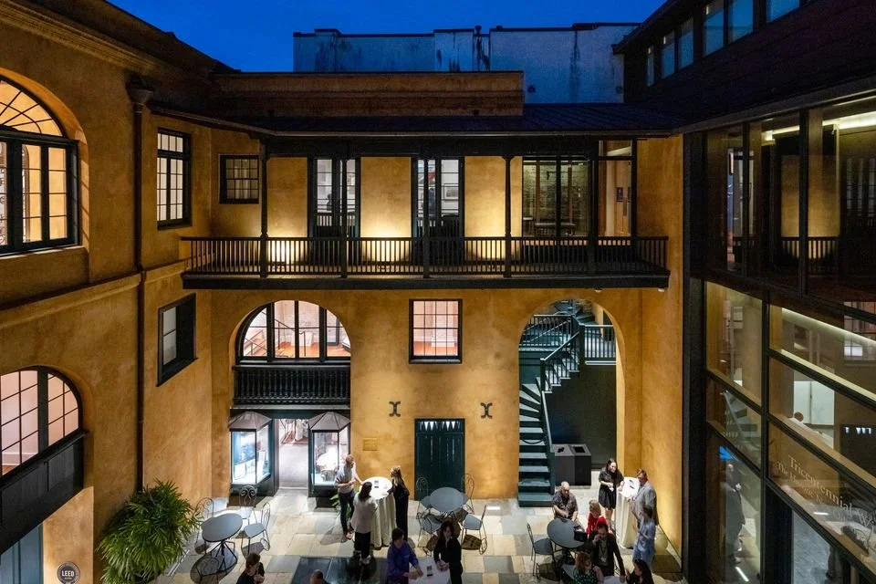 People gather in a historic courtyard with yellow walls, arched doorways, and multiple balconies. The area is illuminated with warm lighting at dusk.