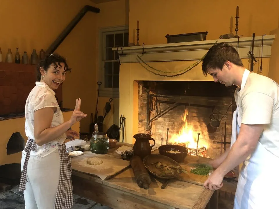 A woman and a man cooking in front of a fireplace. The woman is smiling and making a peace sign with her fingers, while the man is chopping green herbs on a wooden table.