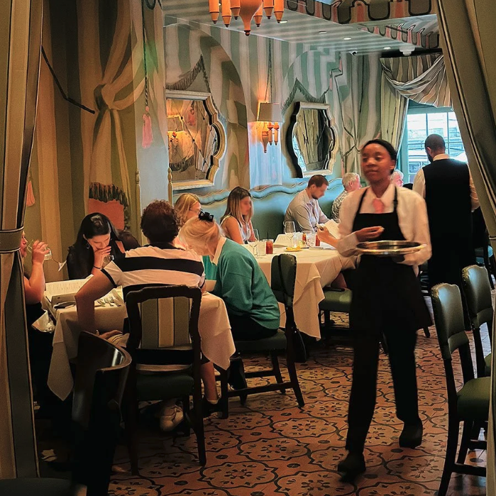 Interior of a restaurant with patrons dining at tables, waitstaff serving food, and green and cream decor with ornate wall mirrors and patterned carpeting.