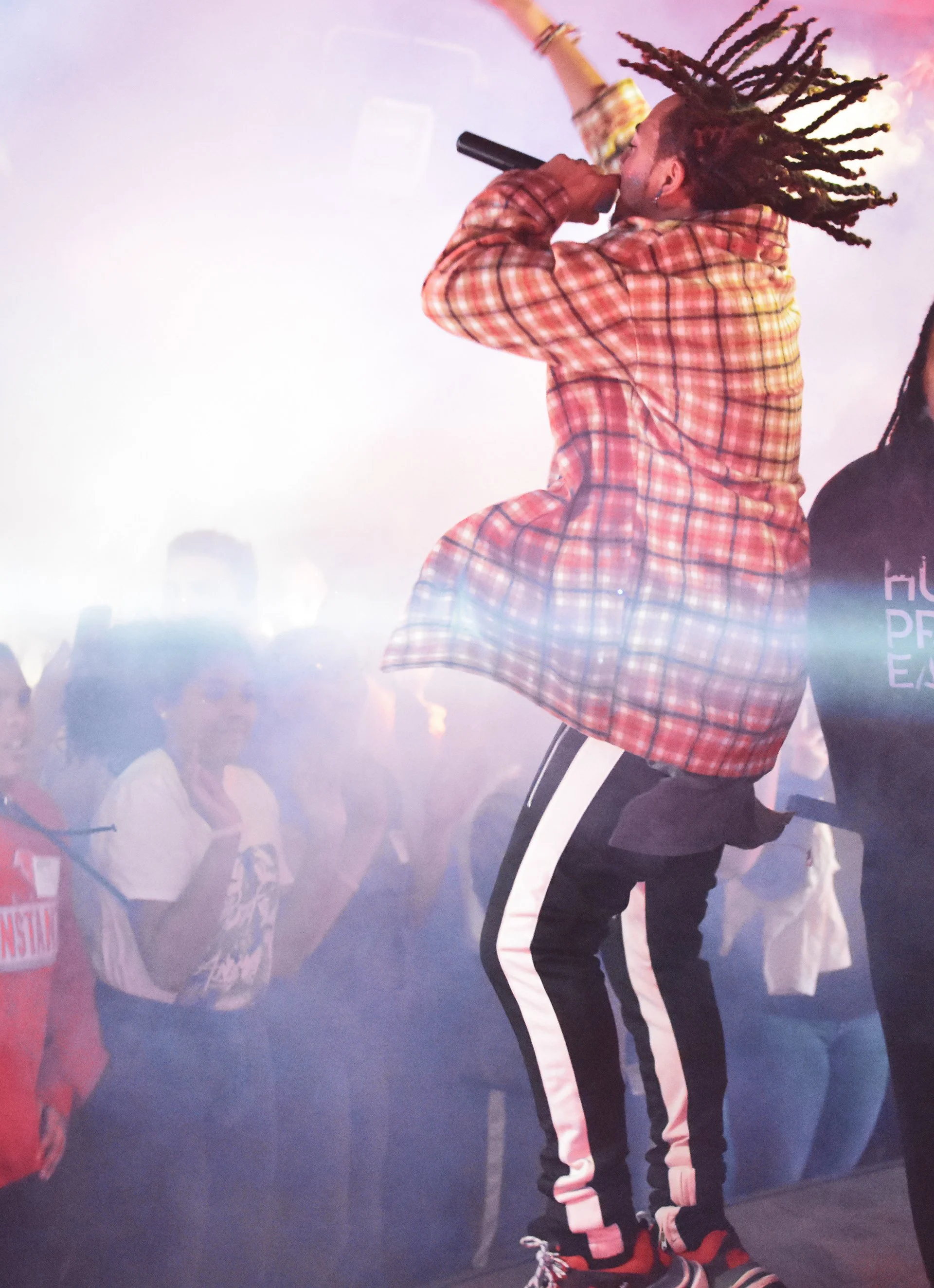Performer on stage with mic, wearing plaid shirt and striped pants, hair in braids, energetic crowd in background