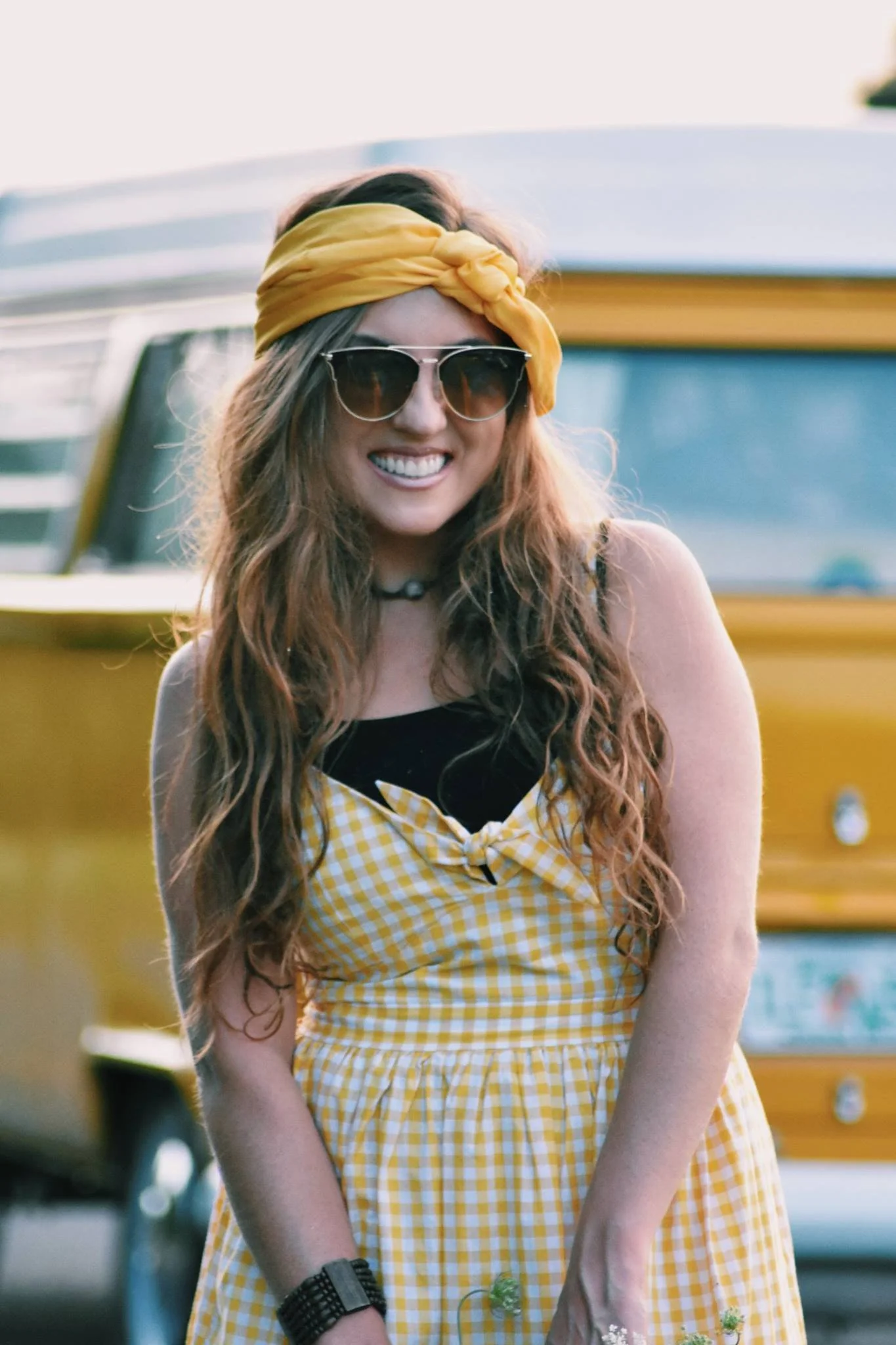 Smiling woman in yellow plaid dress and headband with sunglasses in front of yellow van.