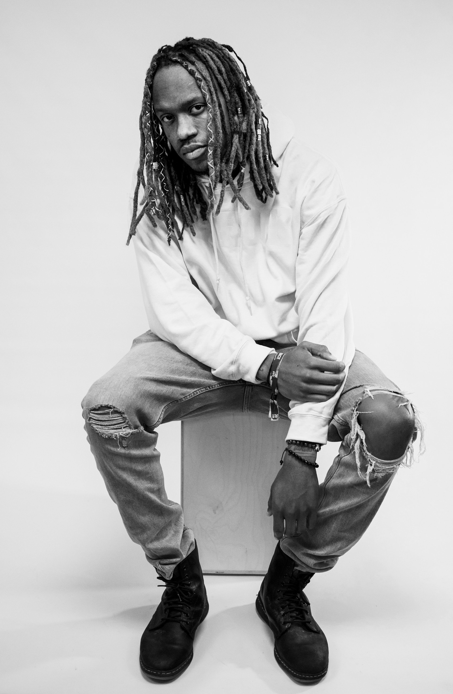 Black and white photo of a person with dreadlocks sitting on a box, wearing a hoodie and ripped jeans, with boots.