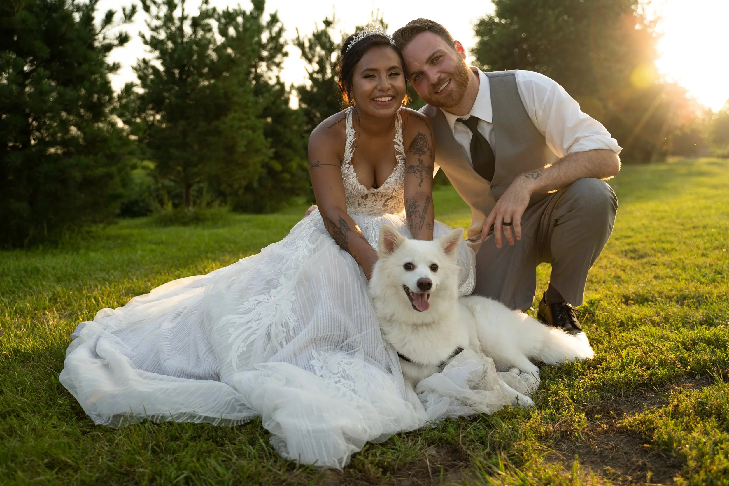 A bride and groom posing outdoors on grass with a white dog, surrounded by trees and sunlight.