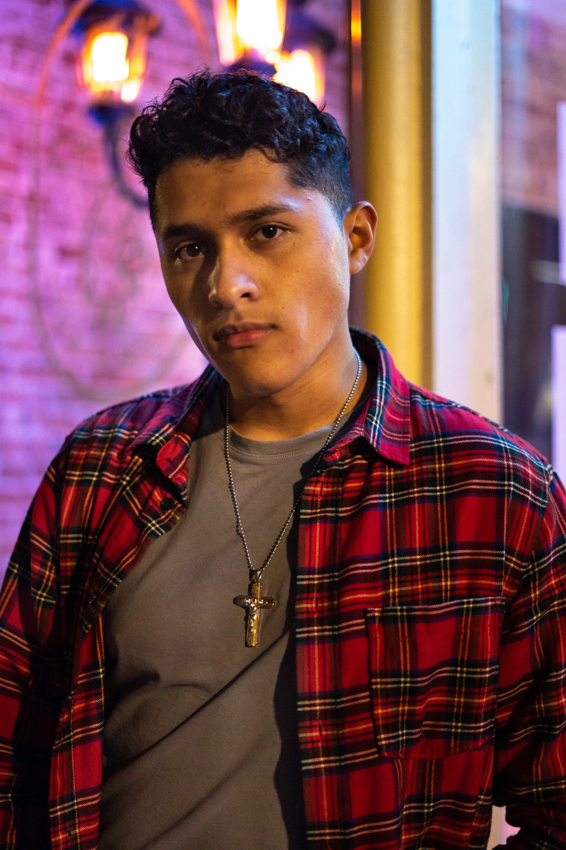 Person wearing a red plaid shirt, gray t-shirt, and cross necklace, standing in front of a blurred, illuminated background.