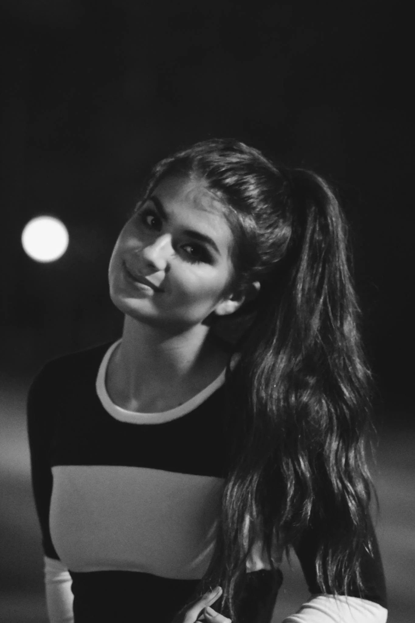 Black and white portrait of a person with a ponytail, wearing a patterned shirt, smiling slightly, with a blurred background.