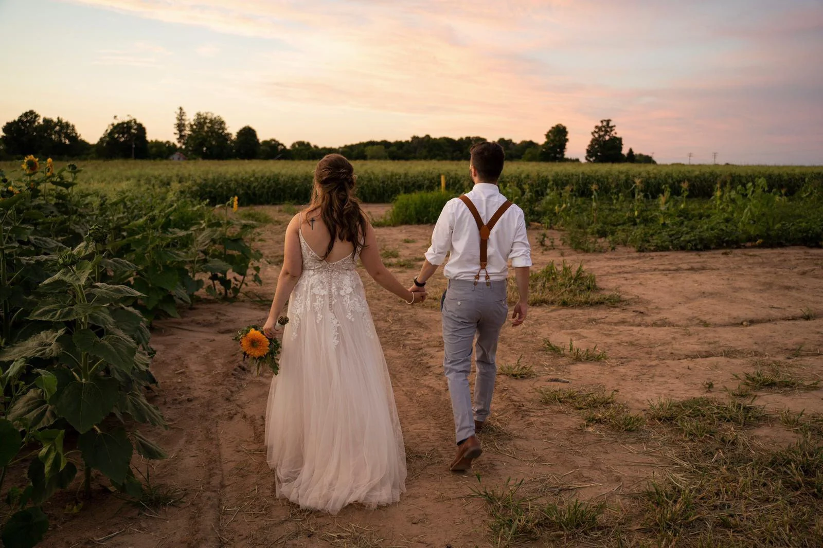 Bride and groom holding hands walking in a field at sunset, with the bride holding a sunflower.