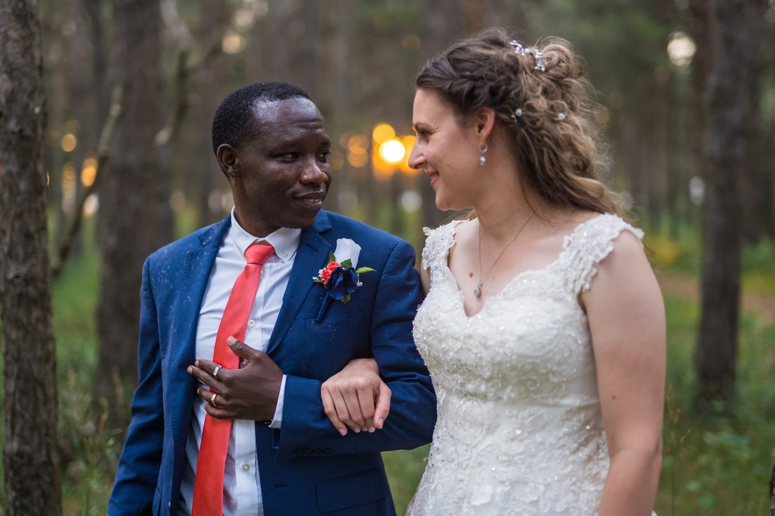 A smiling bride and groom walking in a forest, the groom in a blue suit and red tie, and the bride in a white wedding dress.