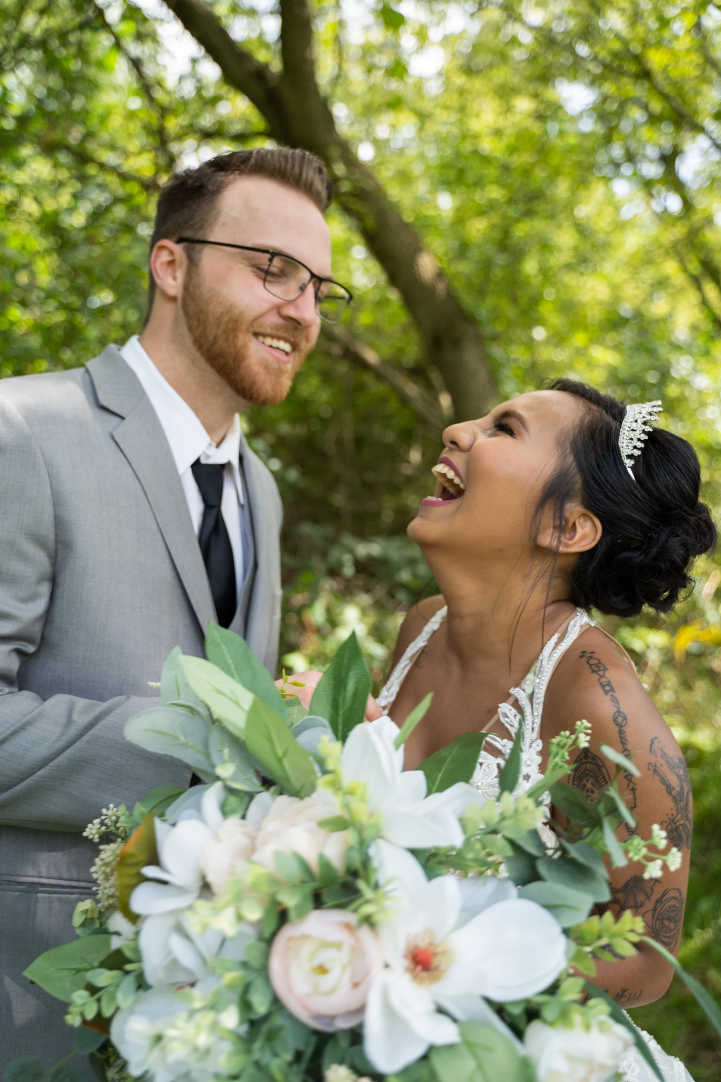 Bride and groom laughing together outdoors with bouquet, surrounded by greenery.