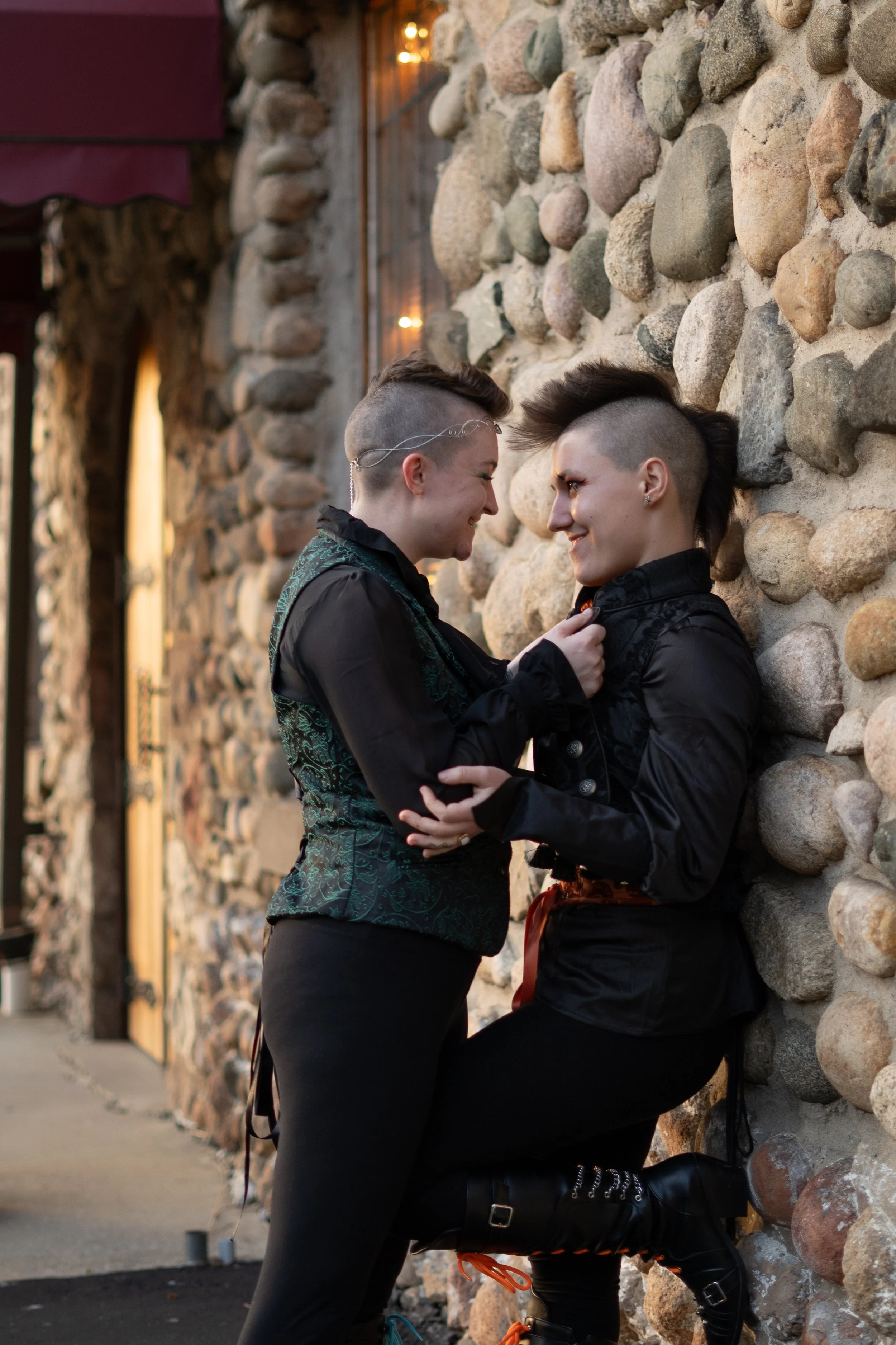Two people with mohawk hairstyles and punk style clothing facing each other closely against a stone wall, smiling and touching.