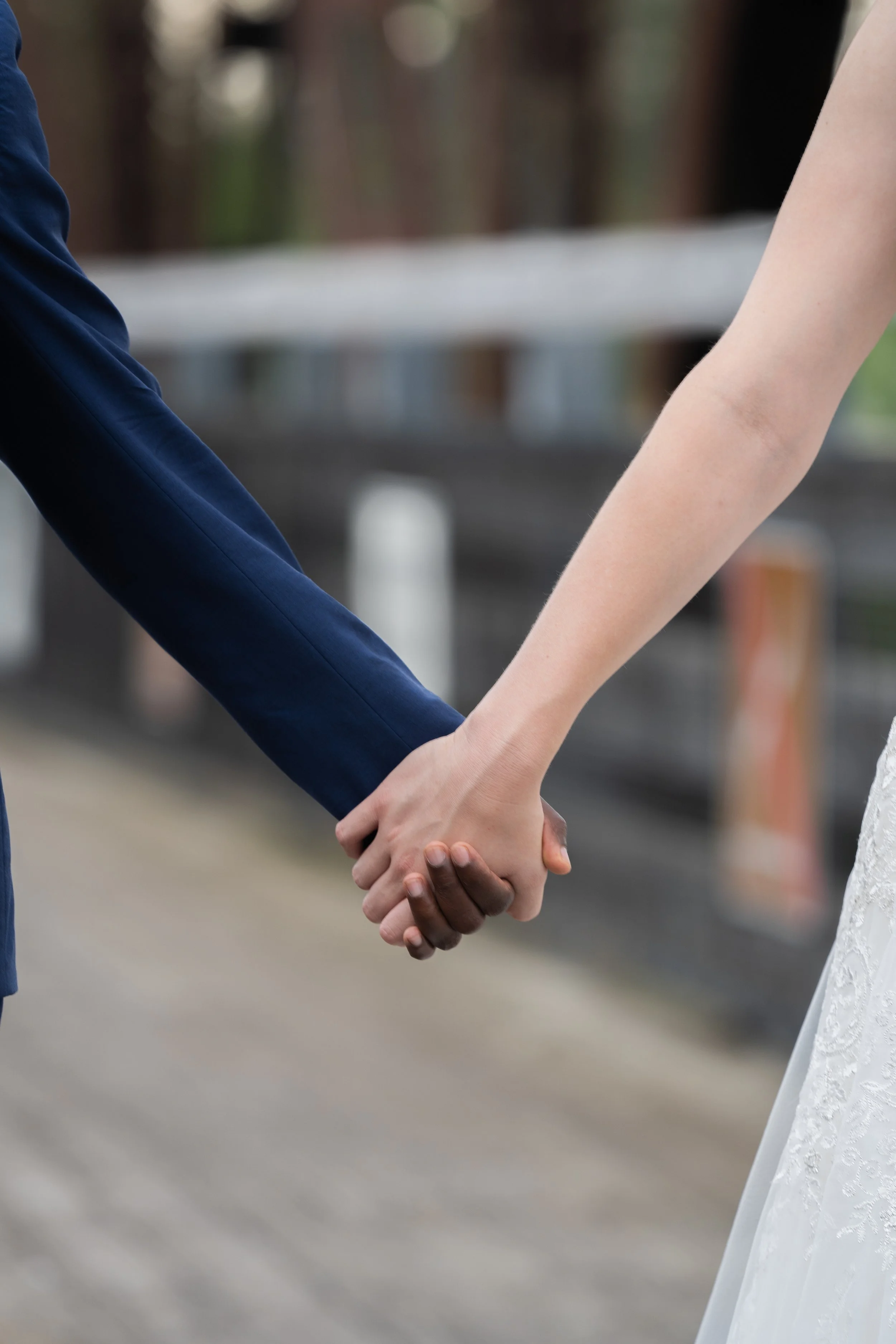 Close-up of a couple holding hands, one in a blue suit and the other in a white dress, likely during a wedding.