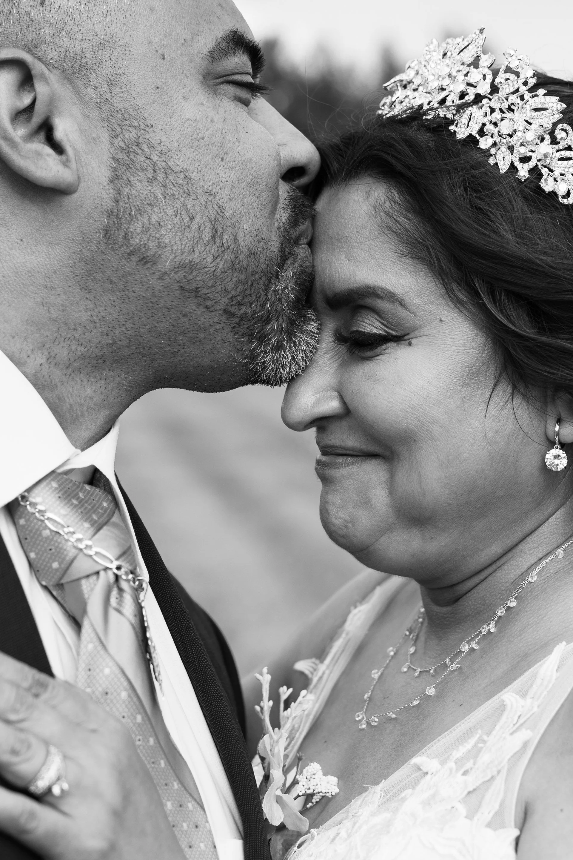 Close-up black and white photo of a groom kissing a bride's forehead, both smiling. The bride wears a tiara and earrings, and the groom is in a suit with a tie.
