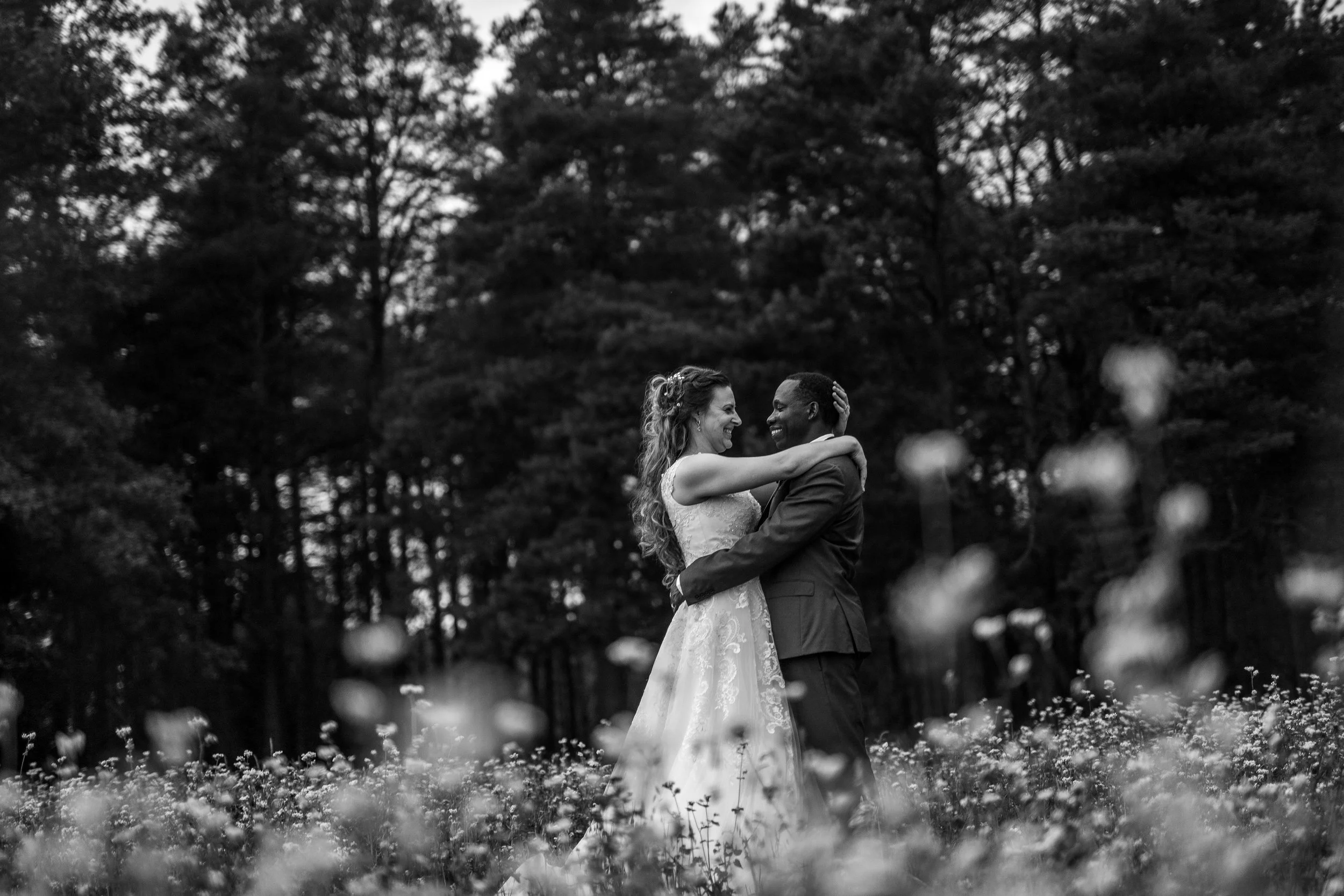 Black and white photo of a couple embracing in a flower field with trees in the background.