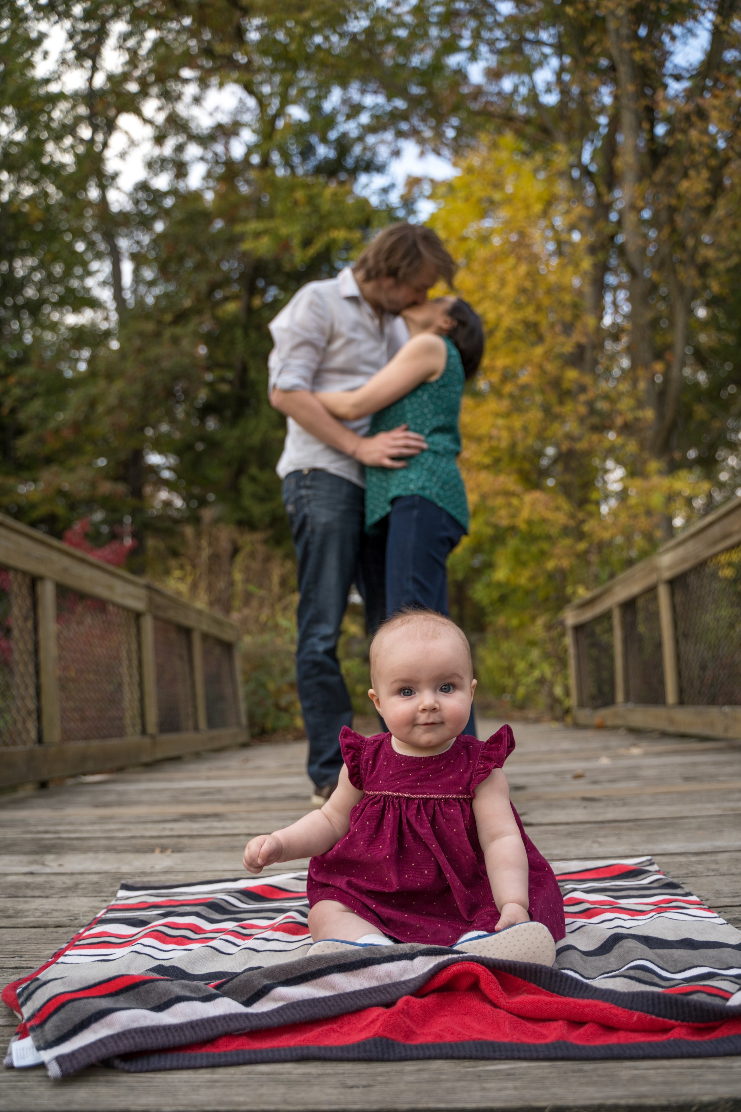 A baby sits on a striped blanket on a wooden walkway with trees in the background. A couple is kissing behind the baby.
