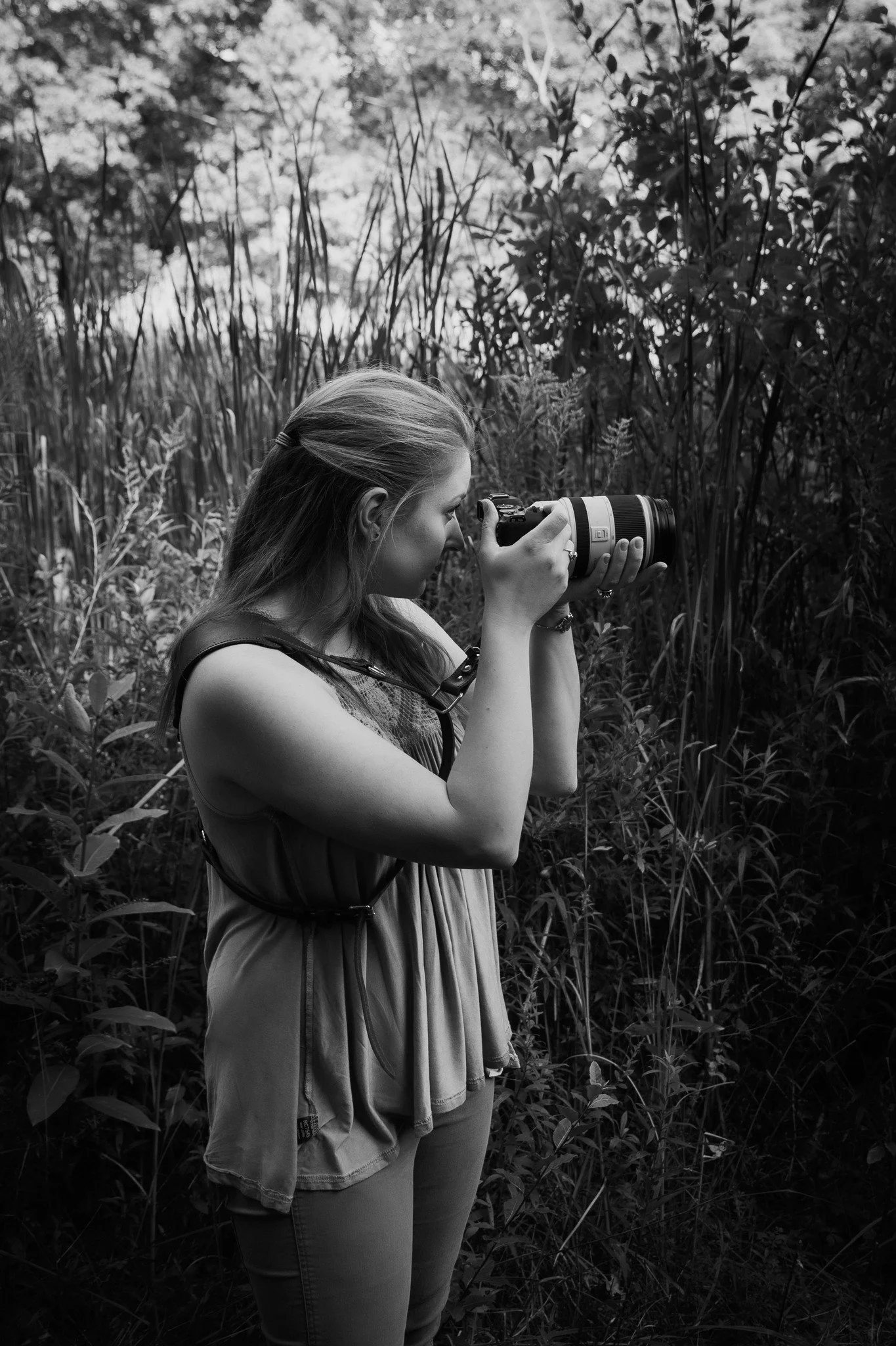 A young woman with long hair taking a photograph with a camera in a natural outdoor setting surrounded by tall grass and bushes.