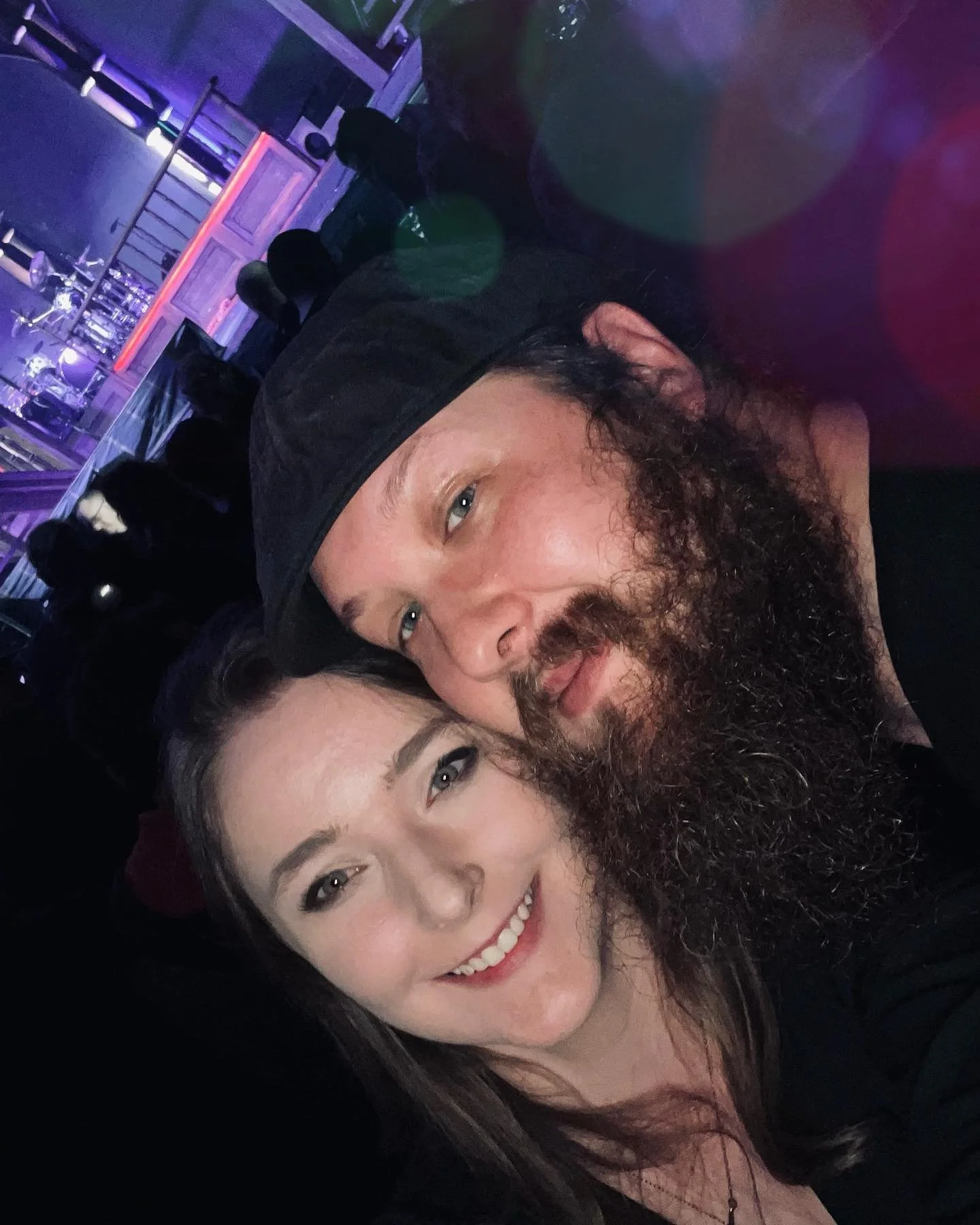 A man with a beard and long hair and woman with long dark hair smiling for a selfie, in a dimly lit place with purple and blue lighting.