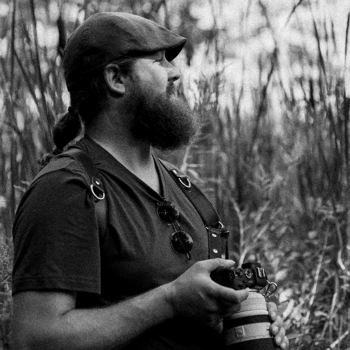 A man with a beard, wearing glasses, a cap, and a backpack, holding a camera in a natural outdoor setting with trees and plants.