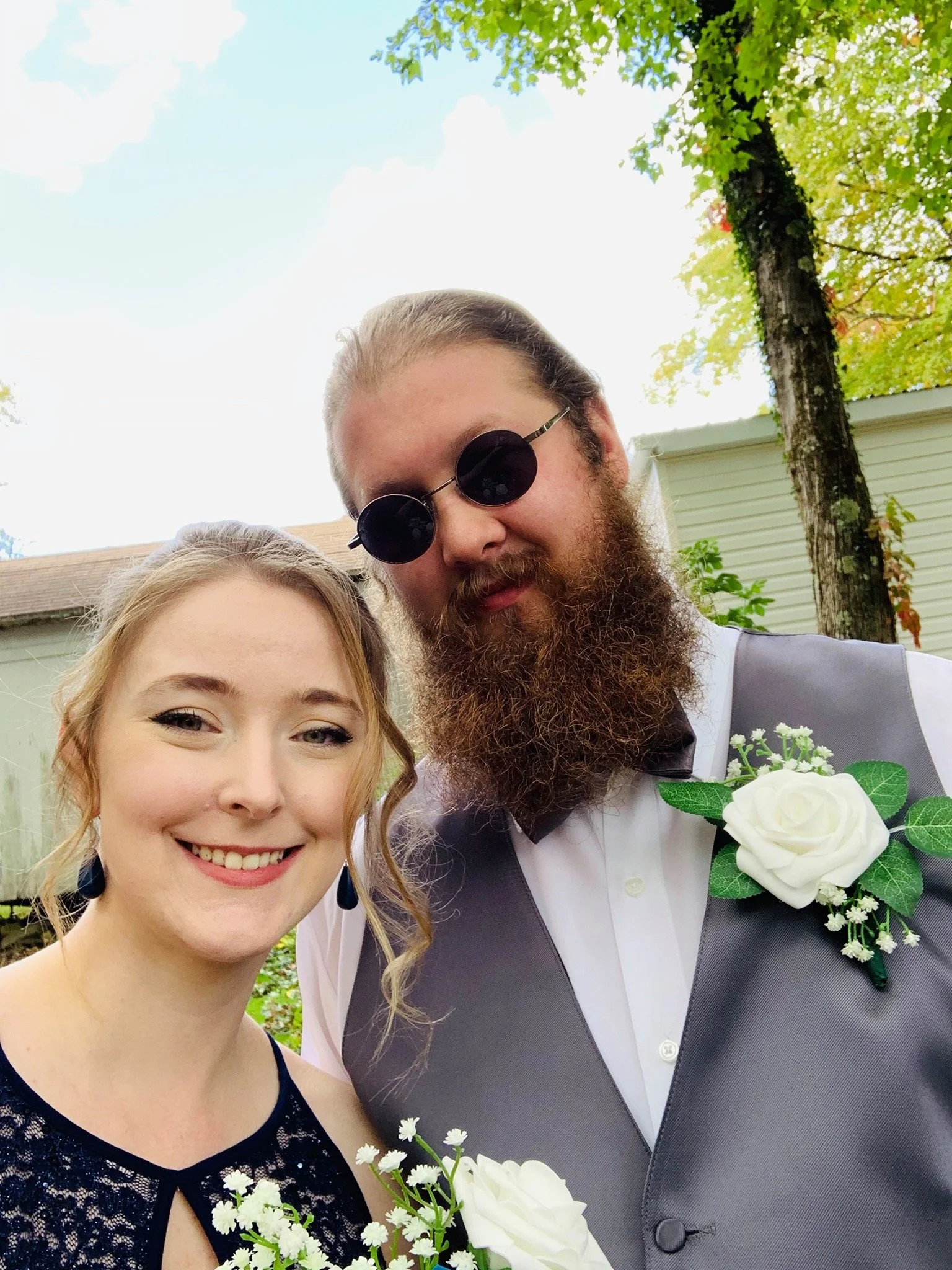 A smiling young woman with curly blonde hair and dark earrings, standing next to a bearded man with sunglasses, both dressed in formal attire, outdoors with trees and a house in the background.