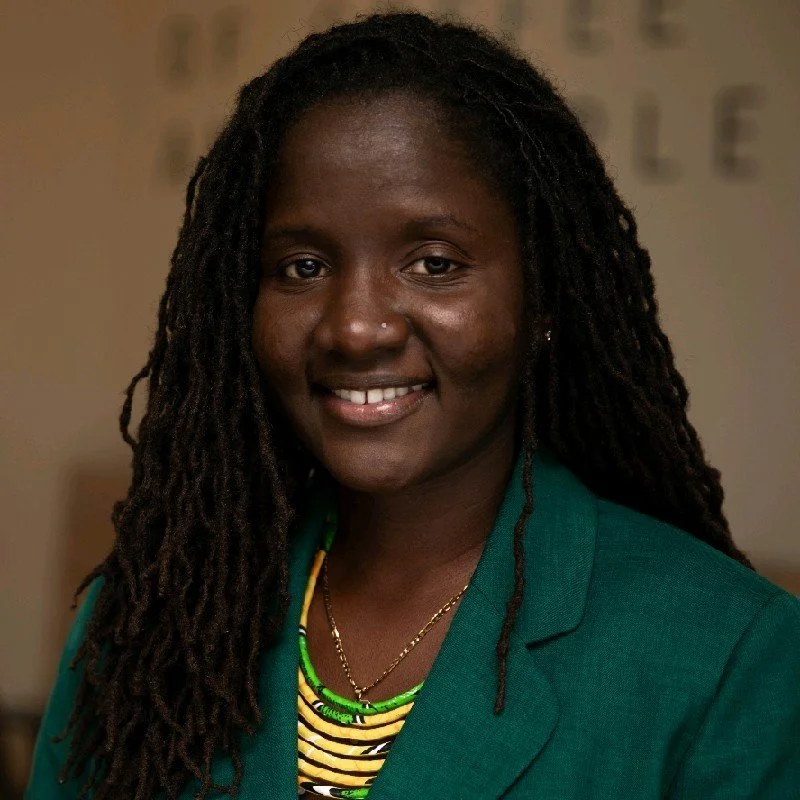 Close-up portrait of a smiling Black woman with long dreadlocks, wearing a green blazer and colorful necklaces.