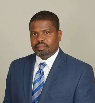 A man in a navy blue suit and striped blue tie posing against a plain light background.