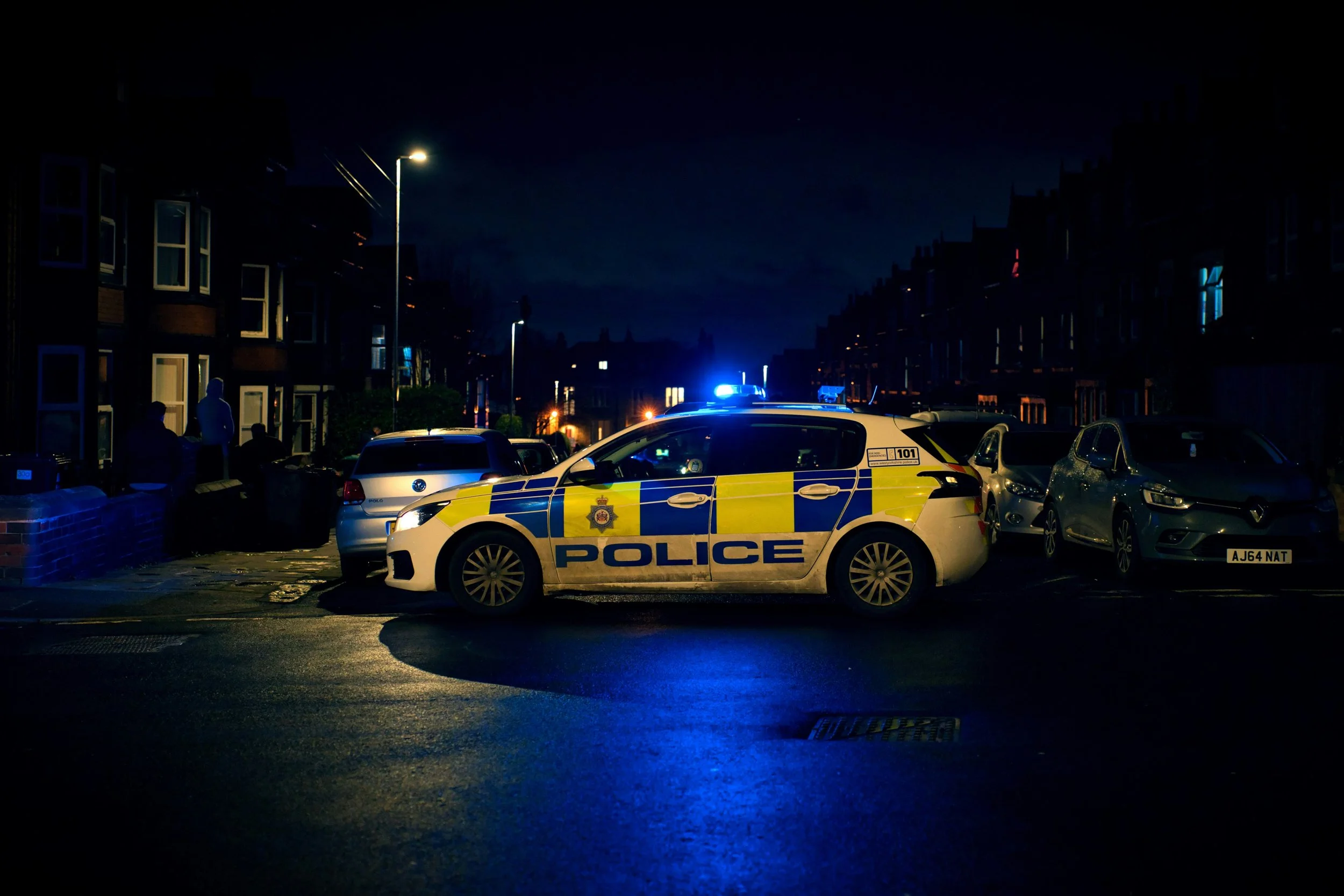 A police car with flashing blue lights parked in a residential neighborhood at night, surrounded by other parked cars and a few people standing nearby.