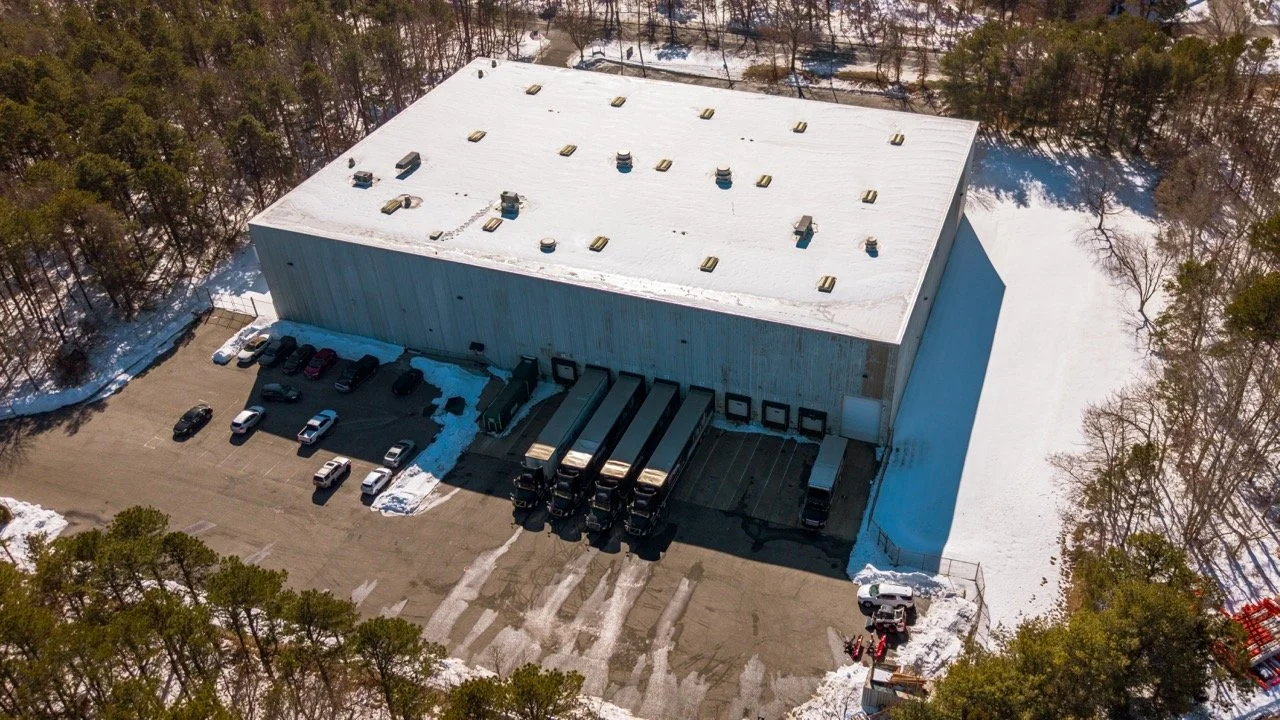 Aerial drone photo of the rear loading docks at 655 Church St in Bayport, NY, with tractor trailers backed into the bays, a parking lot, and snow-covered woods around the warehouse.