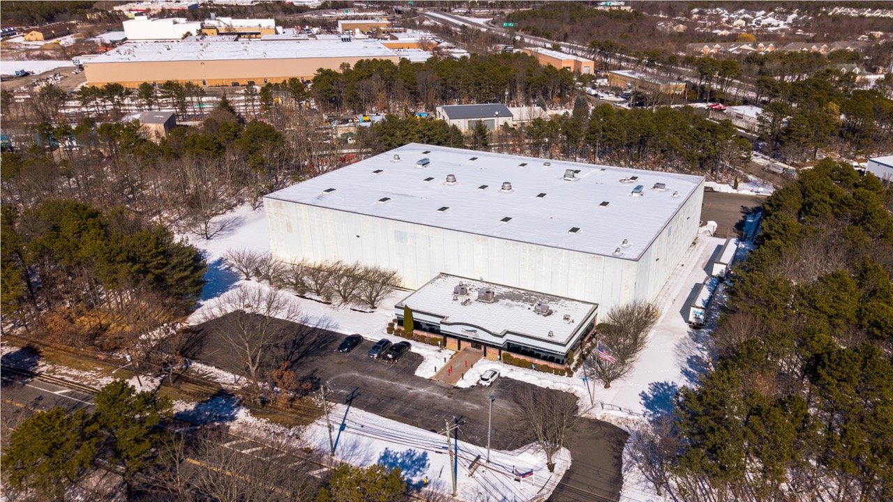Aerial drone photo of the warehouse and front office at 655 Church St in Bayport, NY, surrounded by pine trees and nearby industrial buildings in winter.