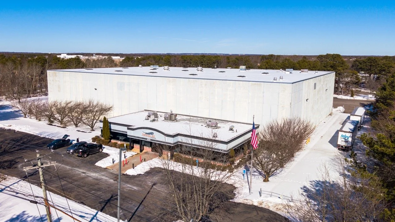 Aerial drone photo of the front entrance and warehouse at 655 Church St in Bayport, NY, with the American flag, parking lot, and trailers along the side in winter.