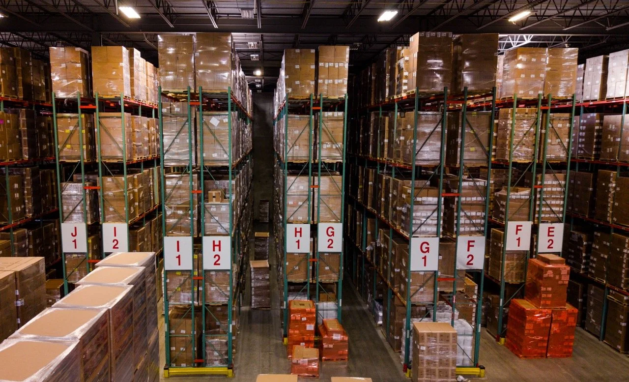 Interior drone photo of the warehouse at 655 Church St in Bayport, NY, showing tall pallet racking with lettered aisle markers from E through J and stacked pallets on the floor.