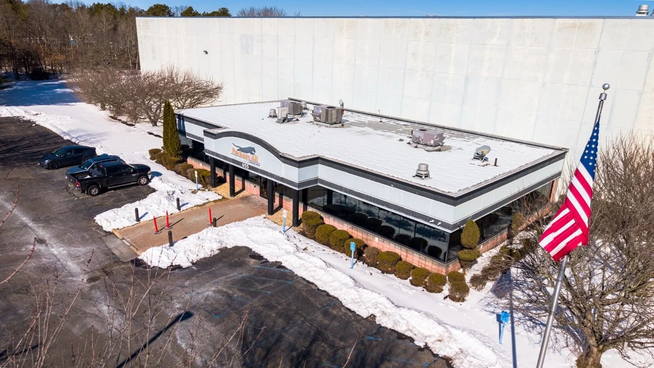 Aerial drone photo of the front office entrance at 655 Church St in Bayport, NY, with the building signage, American flag, and snow-lined walkway in winter.
