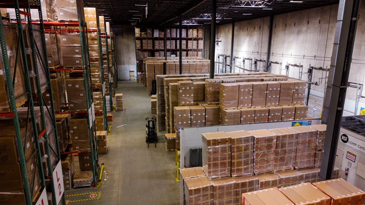 Interior drone photo of the warehouse at 655 Church St in Bayport, NY, looking down a long aisle between pallet racking and rows of stacked, shrink-wrapped pallets.