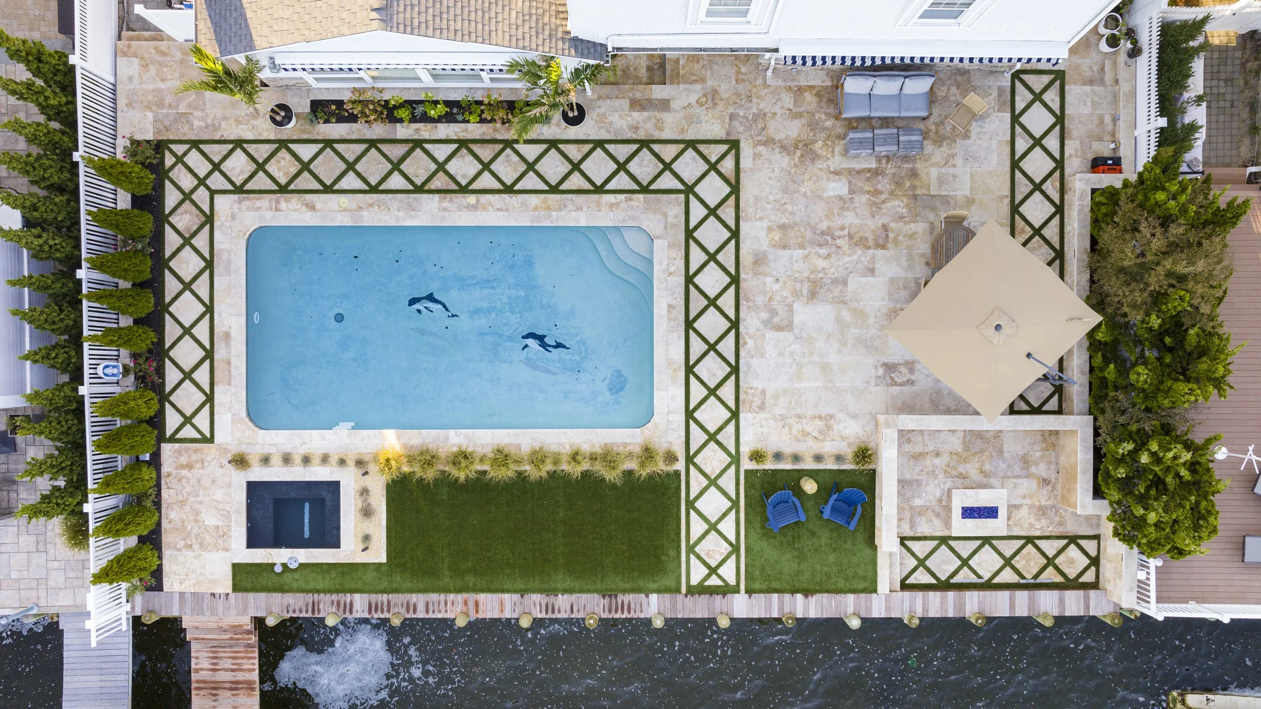 Aerial view of a backyard with a swimming pool, a small hot tub, patio furniture, an umbrella, and landscaped areas bordered by a white fence, with water visible at the bottom edge.