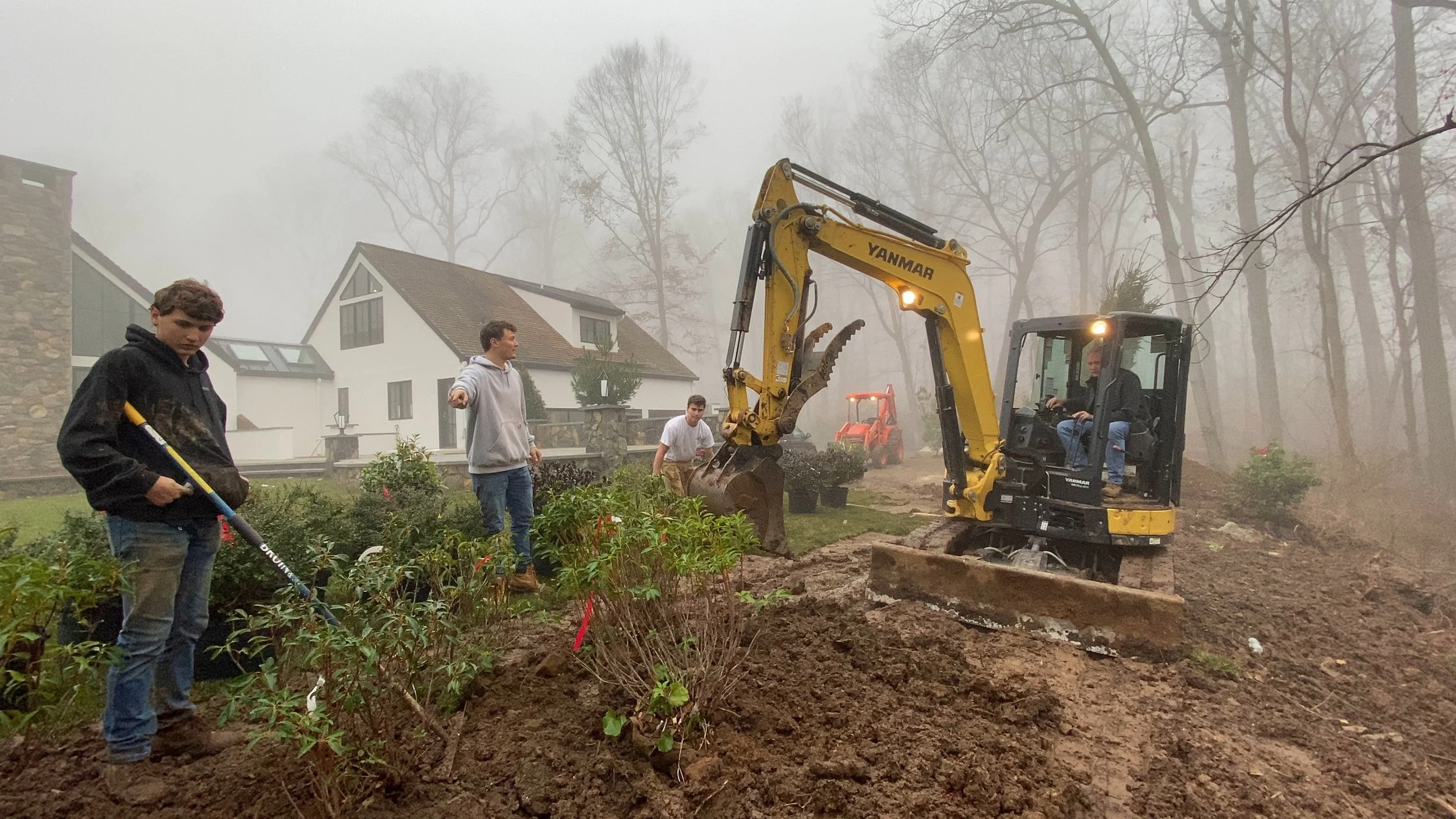 A construction site in a foggy area with a small yellow excavator digging into the ground, three young men are nearby overseeing the work, one holding a rake, with large houses and leafless trees in the background.