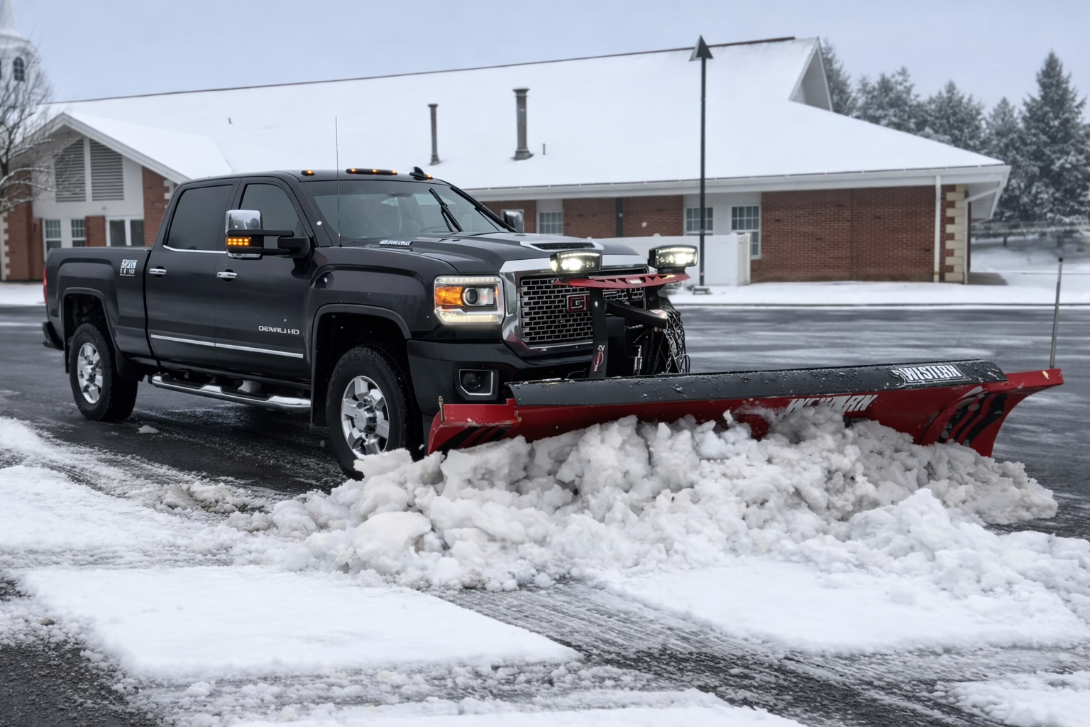 A black pickup truck with a red snow plow attached to the front, clearing snow from a parking lot on a snowy day. In the background, there are snow-covered trees and bushes.