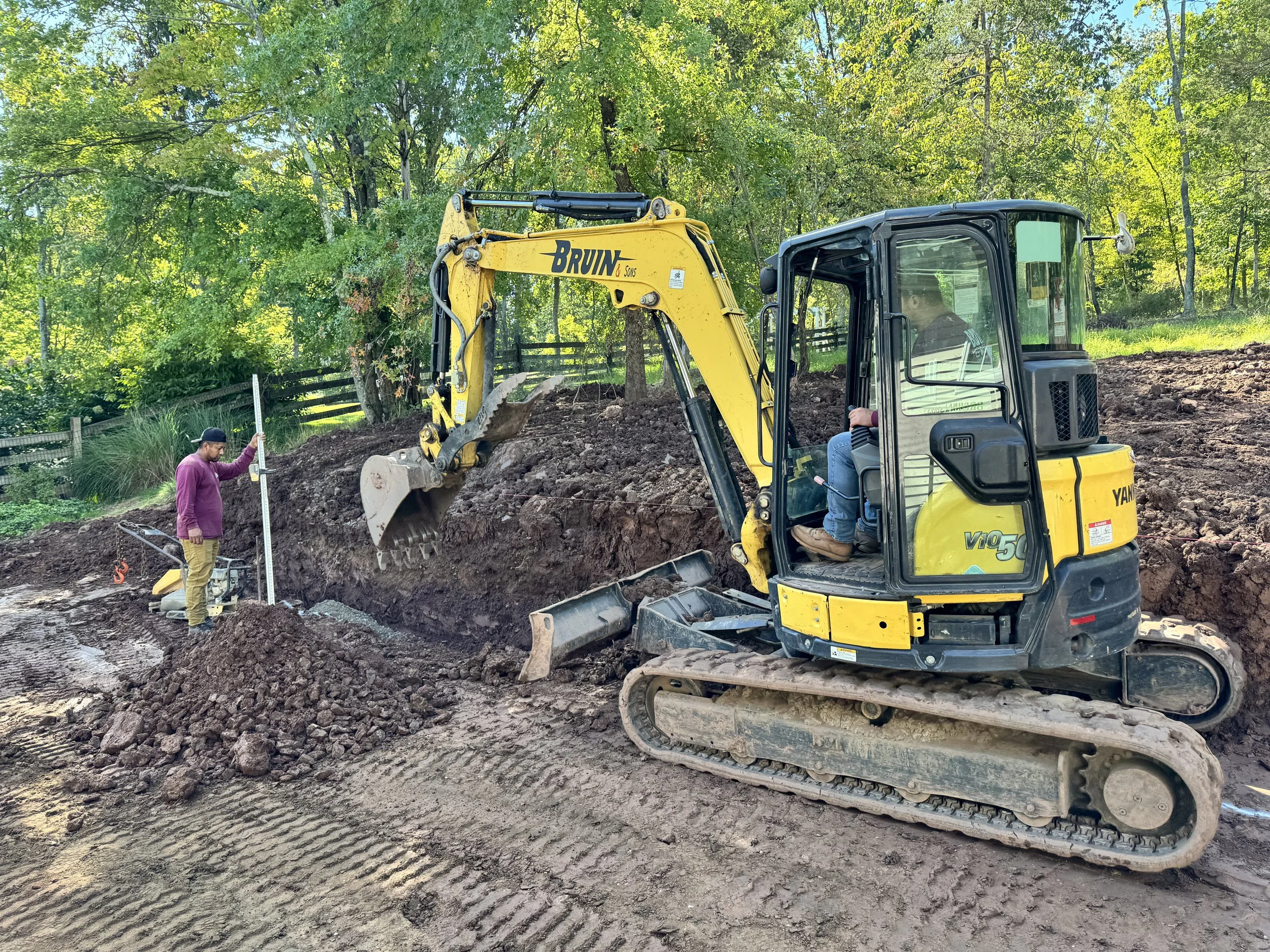 A small yellow excavator demolishing a concrete pool in a backyard with a grassy lawn, trees, and fencing in the background.