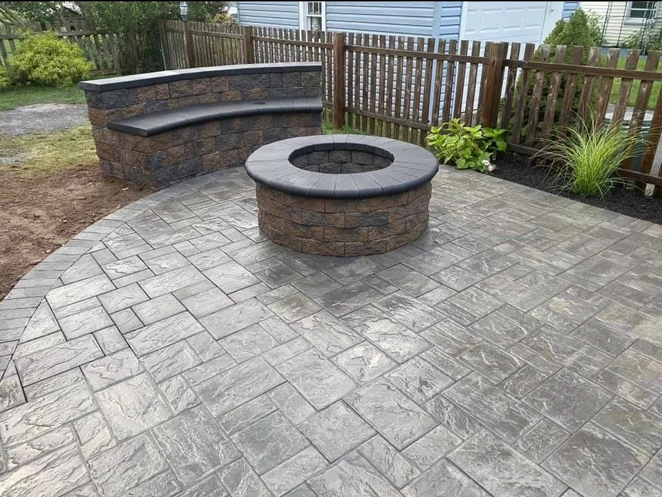 A backyard patio with stamped concrete, a circular fire pit with a stone base, and a curved stone bench with a cushioned top. There is a wooden fence and some plants along the edge.