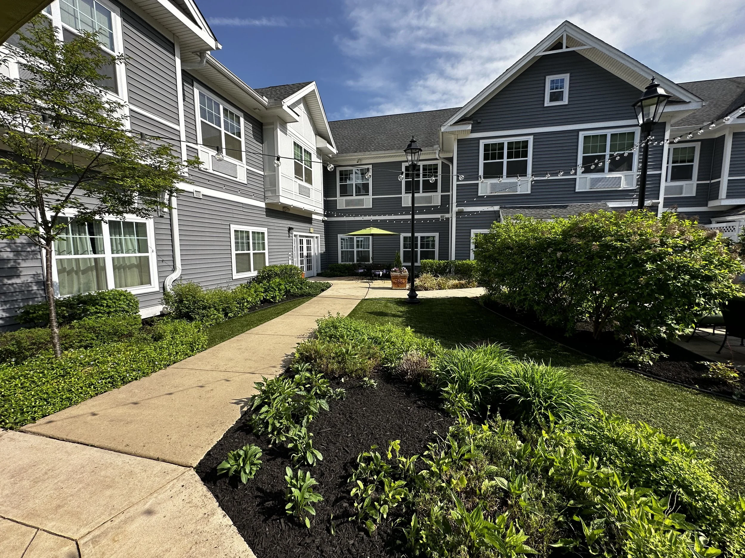 Apartment complex courtyard with landscaped walkway, shrubbery, and outdoor seating under a green umbrella, with string lights and lamp posts.