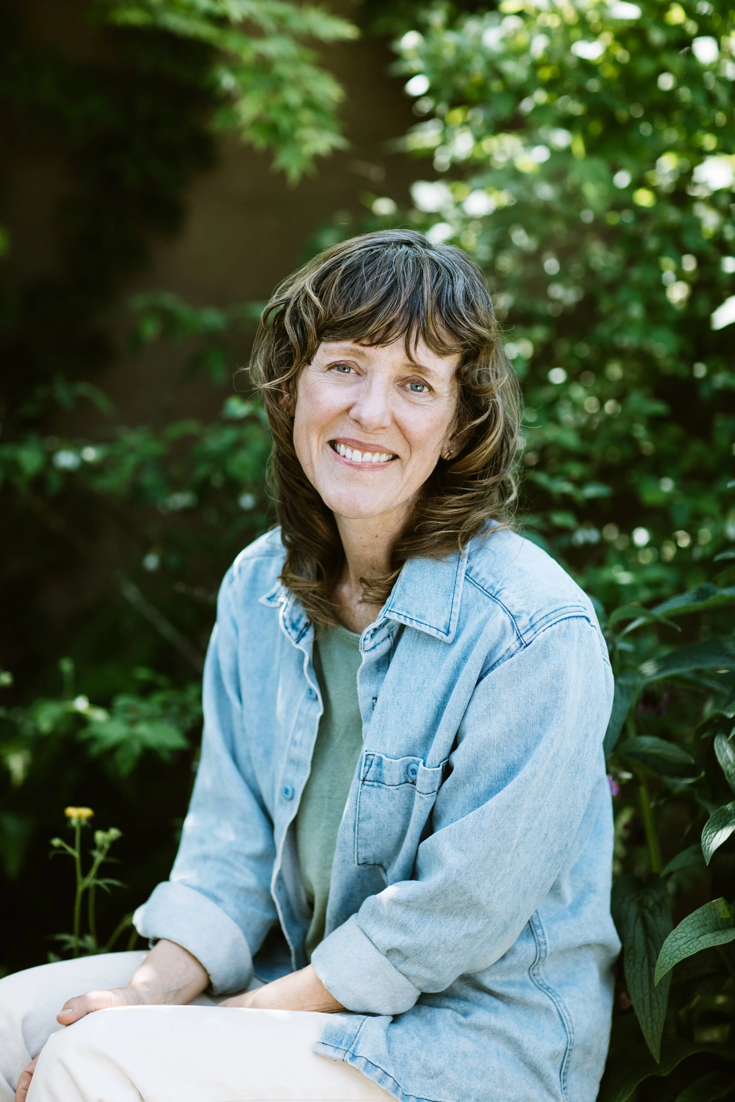 Photograph of a female identifying, white woman in middle age offering spiritual coaching.  She's seated in her permaculture garden and smiling warmly..
