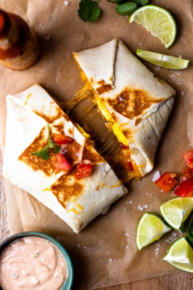 Two burritos on brown parchment paper, with one partially open showing melted cheese and filling, surrounded by lime wedges, cilantro, chopped tomatoes, and a small bowl of pink sauce.
