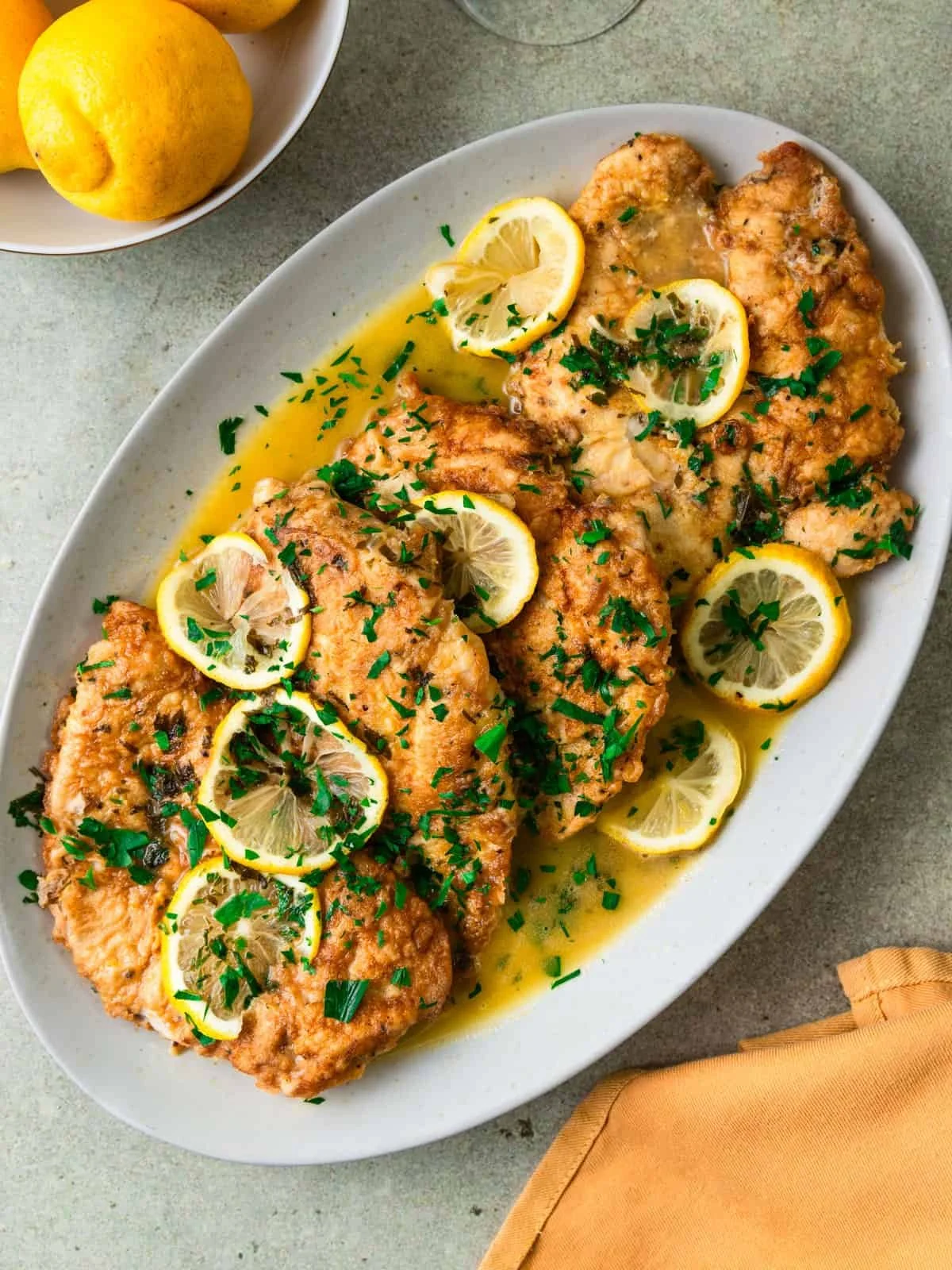 Fried fish fillets topped with lemon slices and chopped herbs, served with lemon wedges and green garnish on a white oval plate.