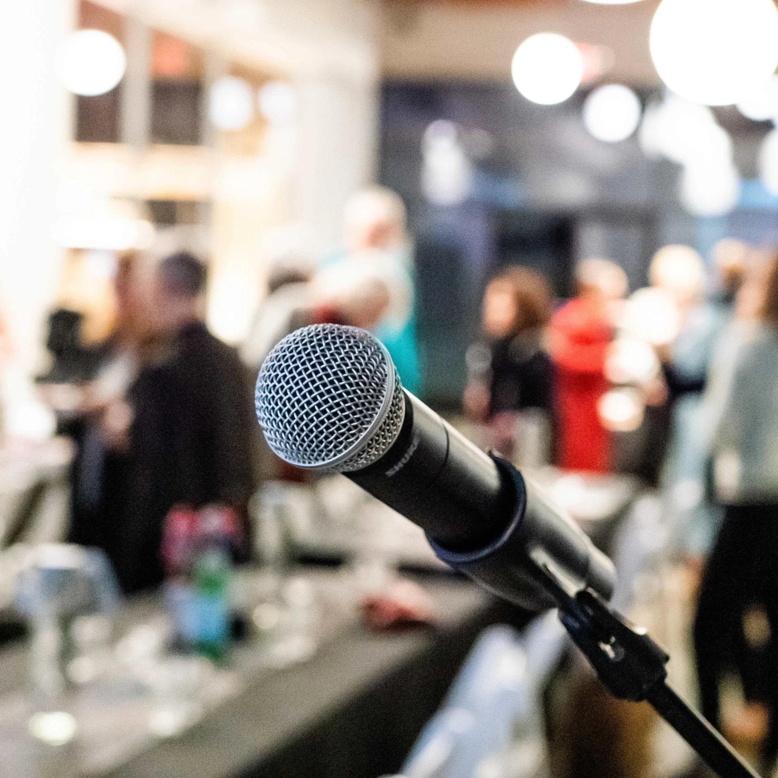 Microphone on a stand in focus with blurred background of people and lights, suggesting a social or event setting.
