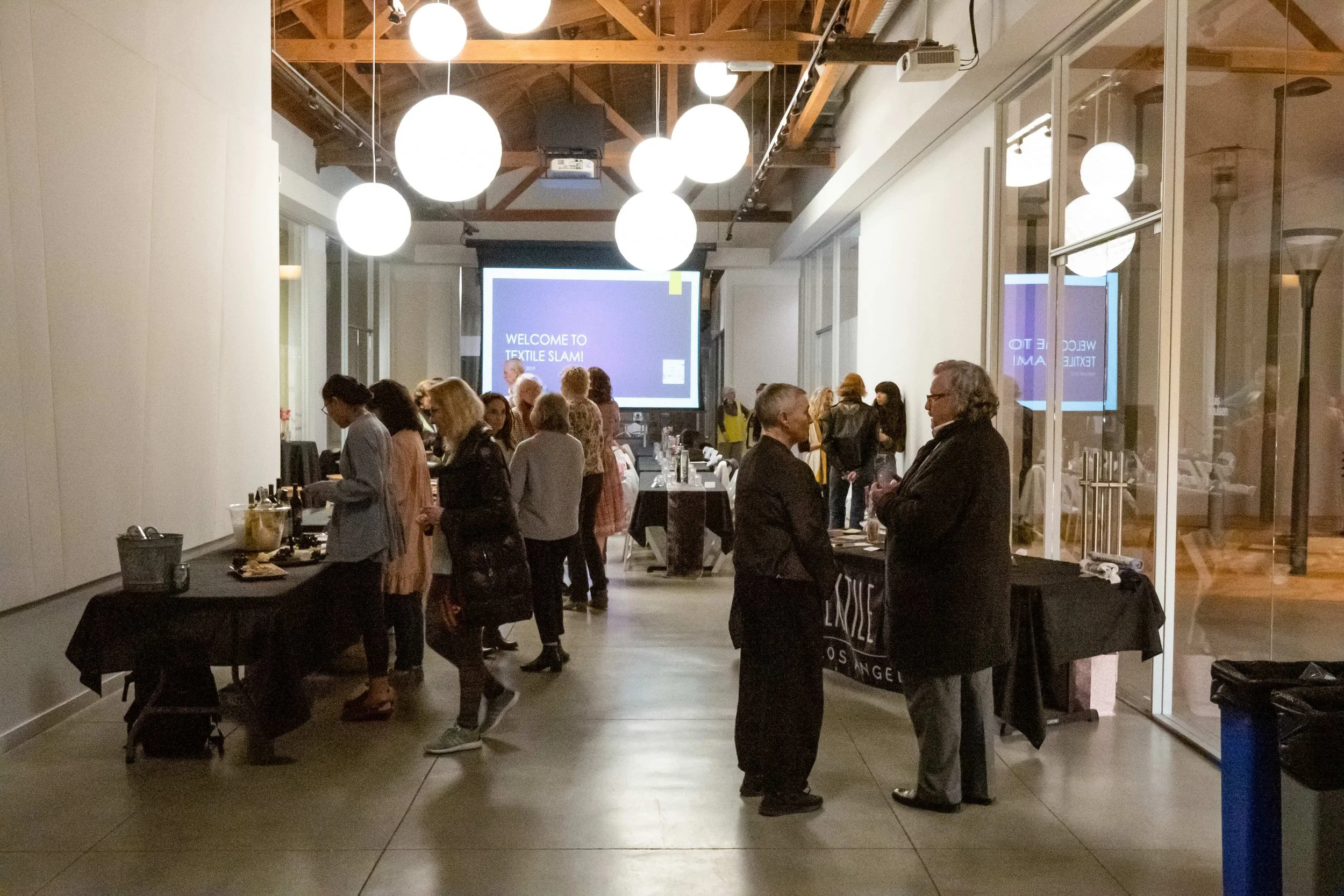 People gathered at an indoor event with a sign reading "Welcome to Textile Slam." Tables with drinks and food are set up, and spherical light fixtures hang from the ceiling.